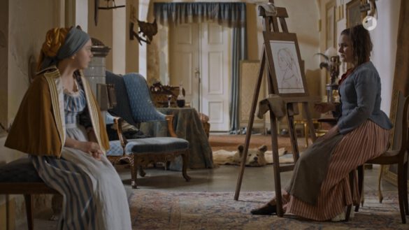 filmstill eines historischen films. in einer schlossartigen situation posiert eine junge Frau für eine junge Malerin an der Staffelei. beide Personen sitzen.