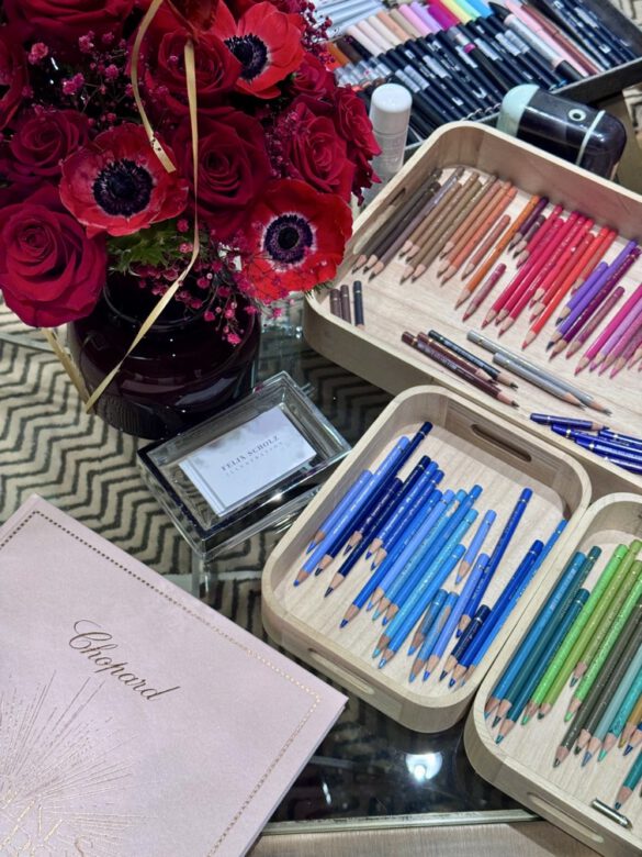 Farbige Zeichenutensilien in Tabletts auf einem Glastisch mit roten Blumen in einer Vase, dazu eine Mappe mit goldenem Chopard Logo