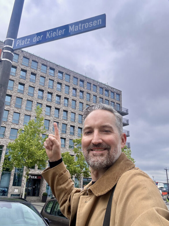 Der Künstler Felix Scholz zeigt auf das Kieler Straßenschild Platz der Kieler Matrosen. Im Hintergrund ein modernes Gebäude